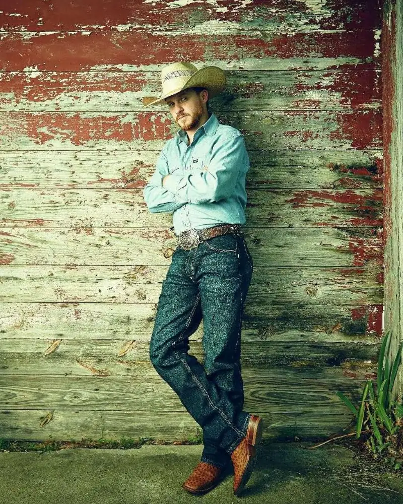 Cody Johnson in a straw cowboy hat, leaning against a weathered red barn door with his arms crossed