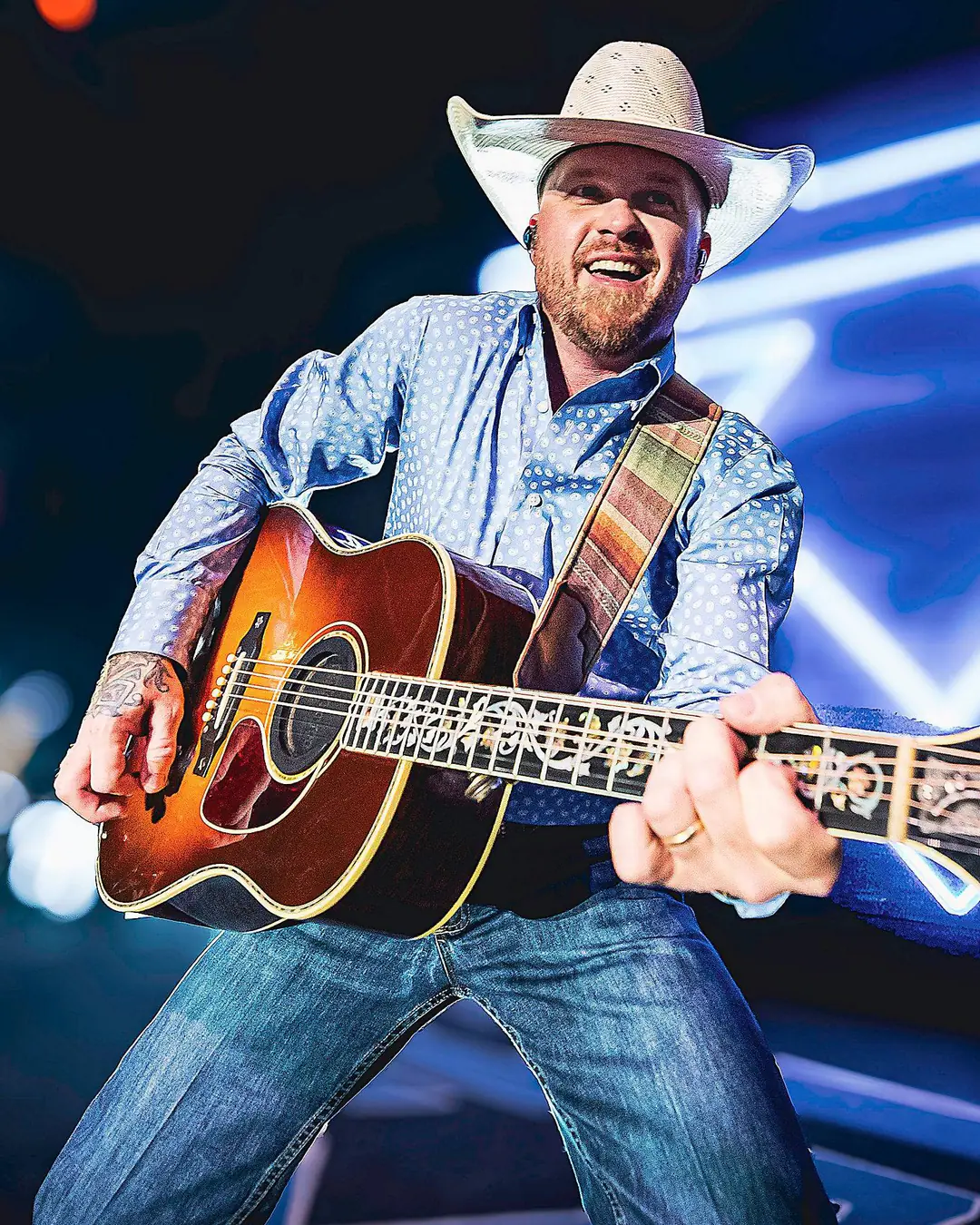 Cody Johnson smiling on stage, playing an acoustic guitar under blue stage lights